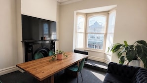 A living room with a wooden dining table, green chairs, a black sofa, a flat-screen TV, a washing machine, and a large window with a view of a building. - Brunsi hostel (Brunswick)