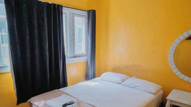 A single bed with a white mattress and a beige blanket, a small bedside table, a round mirror, and a window with curtains. - Clarendon Hotel (Huntington Park)