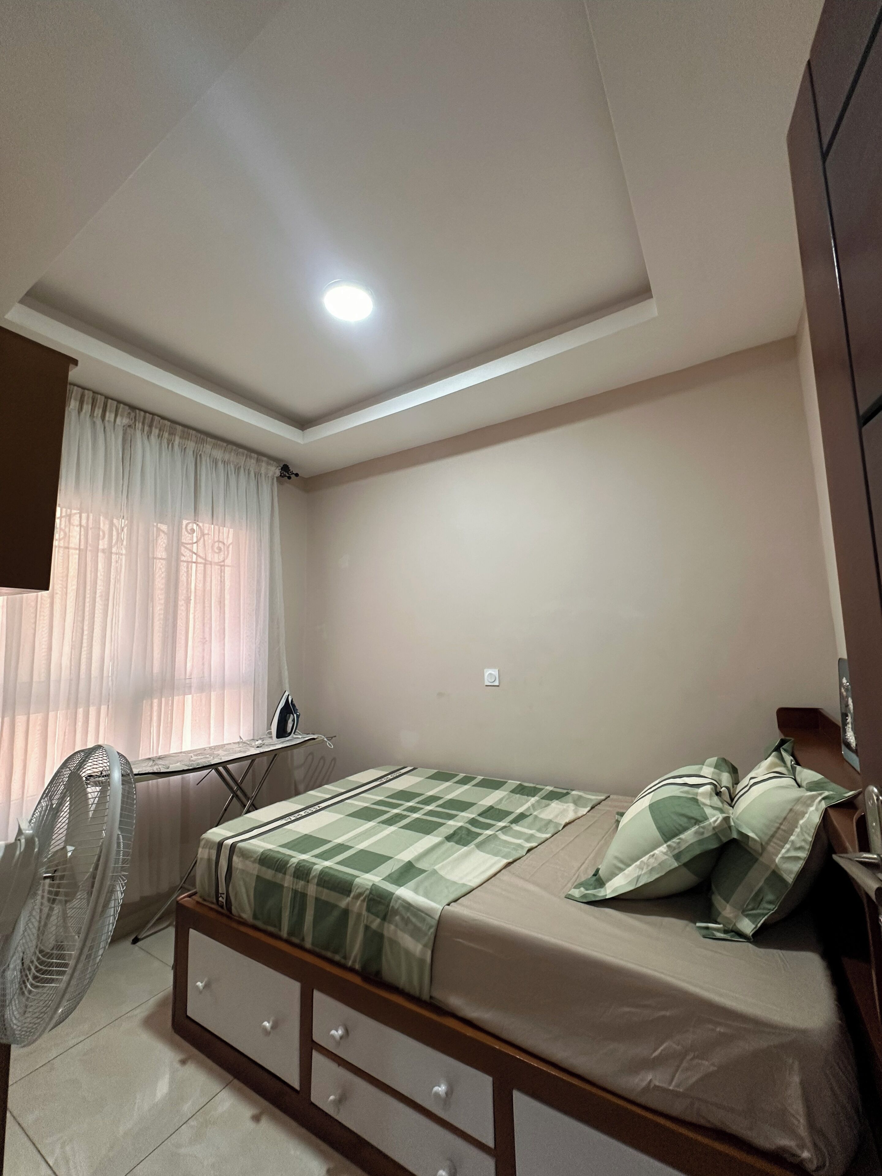 Een slaapkamer met een bed, een bureau, een stoel, een ventilator en een raam met gordijnen.