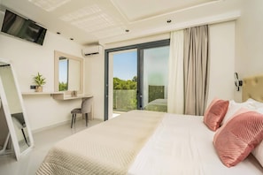 Ein modernes Schlafzimmer mit einem großen Bett, einem an der Wand befestigten Fernseher, einem Schreibtisch mit Stuhl und einer Schiebeglastür, die auf einen Balkon führt.