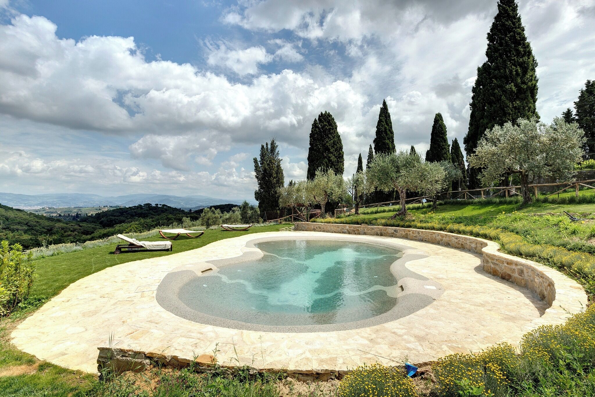 Une piscine circulaire bordée de pierres, entourée d’une pelouse avec des arbres et offrant une vue lointaine sur des collines.