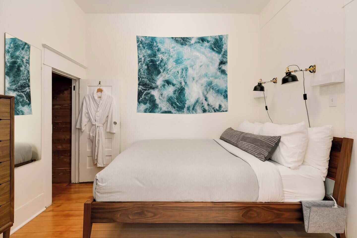 A bedroom with a wooden bed, white bedding, a large abstract painting, a robe hanging on a door, and a bedside lamp.