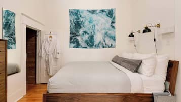 A bedroom with a wooden bed, white bedding, a large abstract painting, a robe hanging on a door, and a bedside lamp.