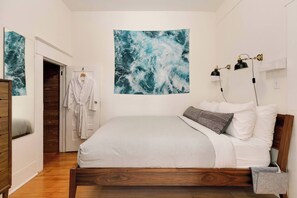 A bedroom with a wooden bed, white bedding, a large abstract painting, a robe hanging on a door, and a bedside lamp. - Cozy Georgetown Apartment (Seattle)