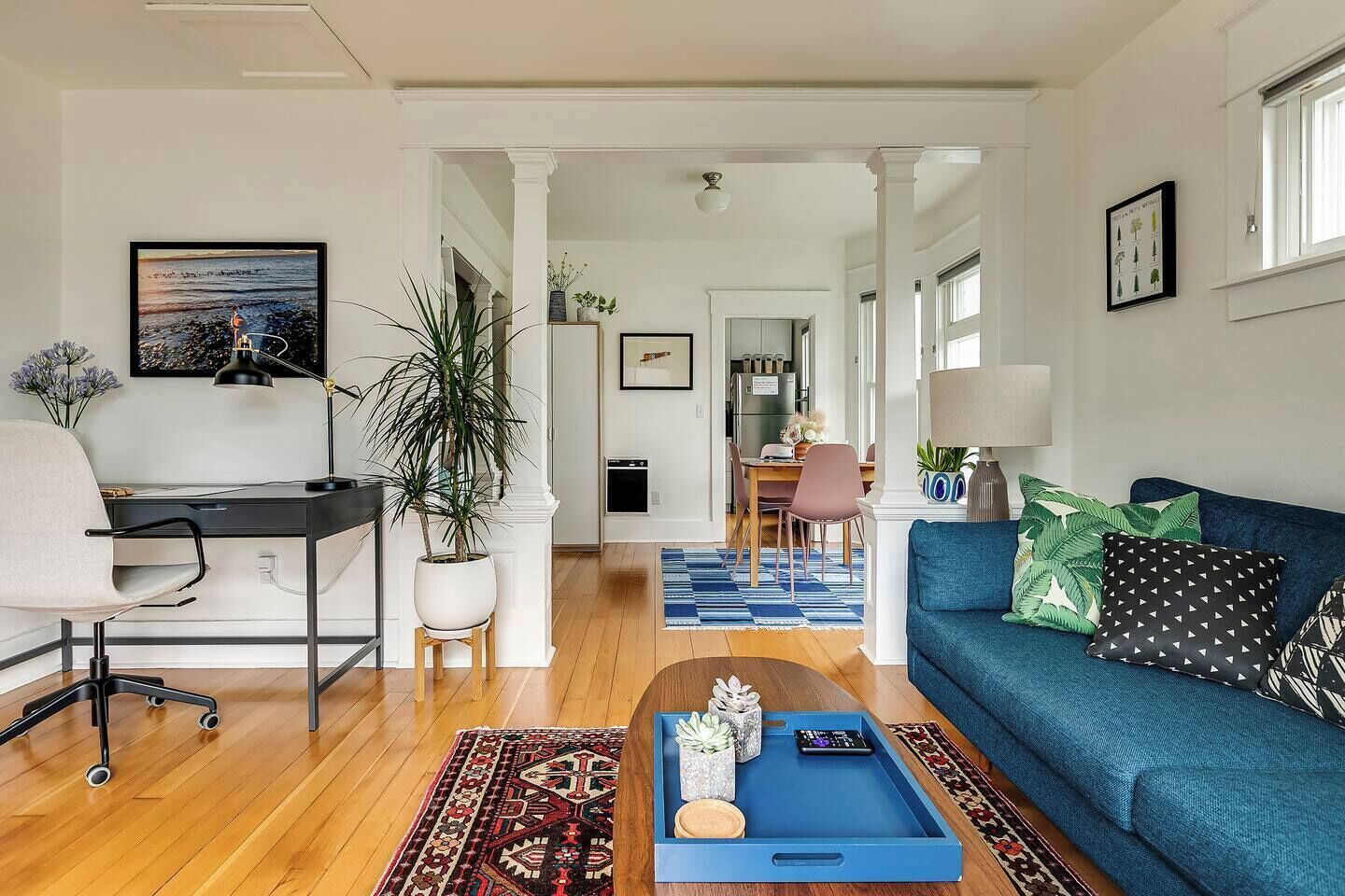 A modern living room with a blue sofa, a desk with a chair, a coffee table with decorative items, and a dining area in the background.
