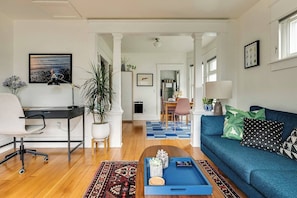 A modern living room with a blue sofa, a desk with a chair, a coffee table with decorative items, and a dining area in the background. - Cozy Georgetown Apartment (Seattle)