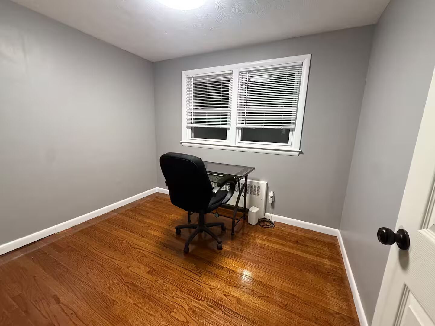 A room with a wooden floor, a black office chair, a white radiator, and a window with blinds.