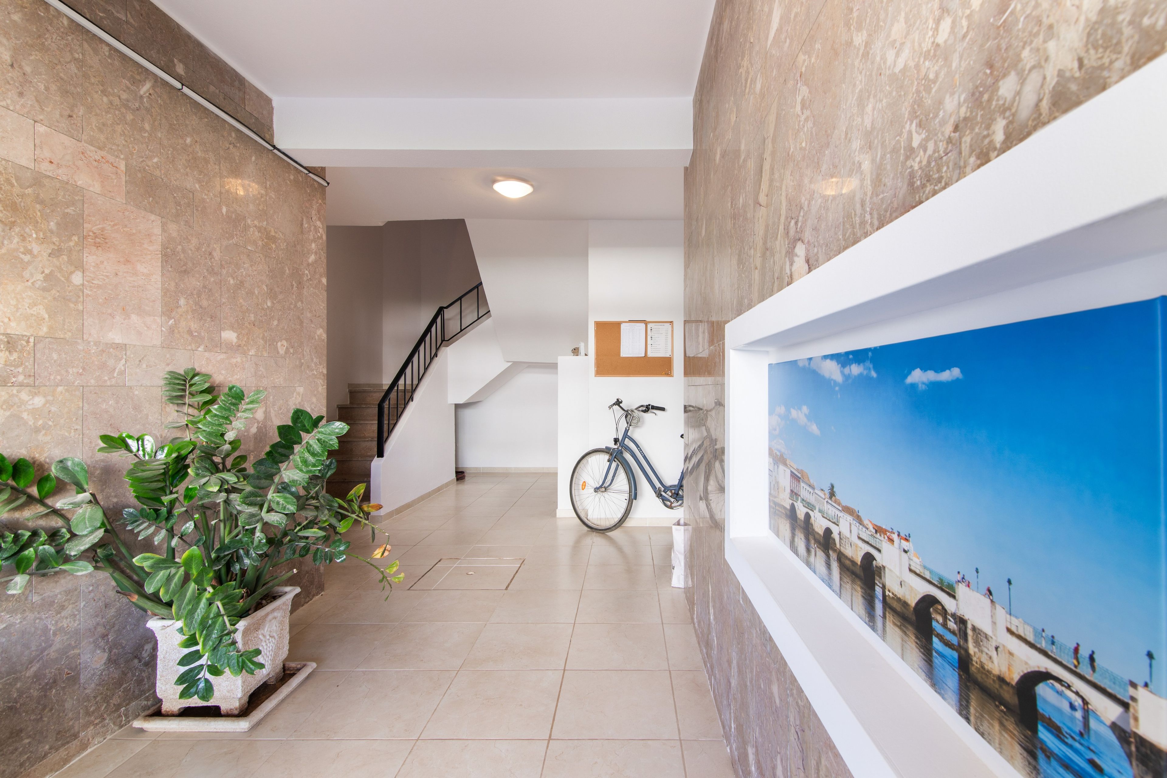 A hallway with a staircase, a potted plant, a bicycle, and a mural of a bridge.