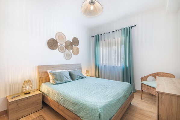 Une chambre à coucher avec un lit en bois, une table de chevet, une lampe, une chaise en osier et un luminaire fixé au mur.