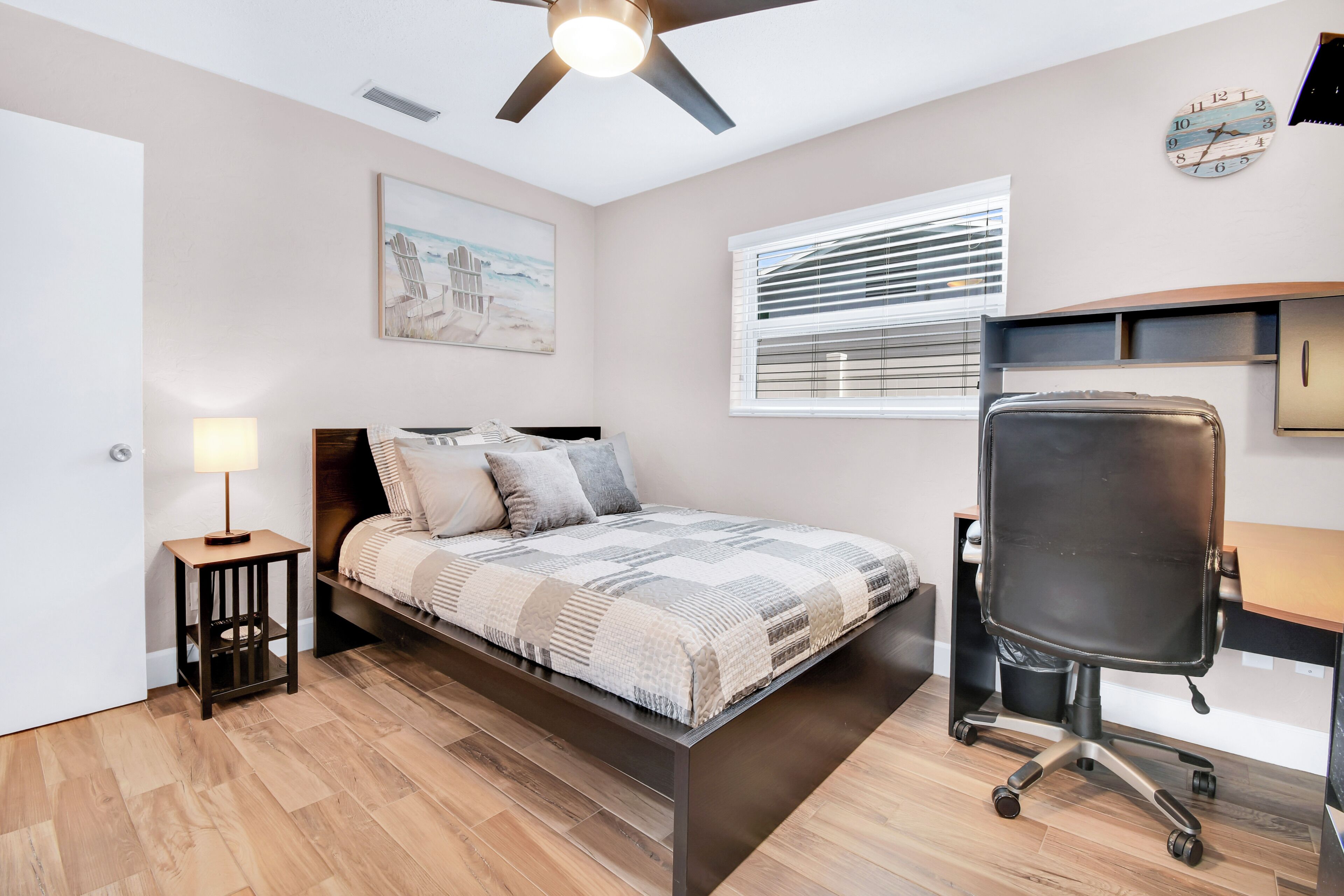 A bedroom with a bed, a desk, a chair, a ceiling fan, a window with blinds, and a wall clock.