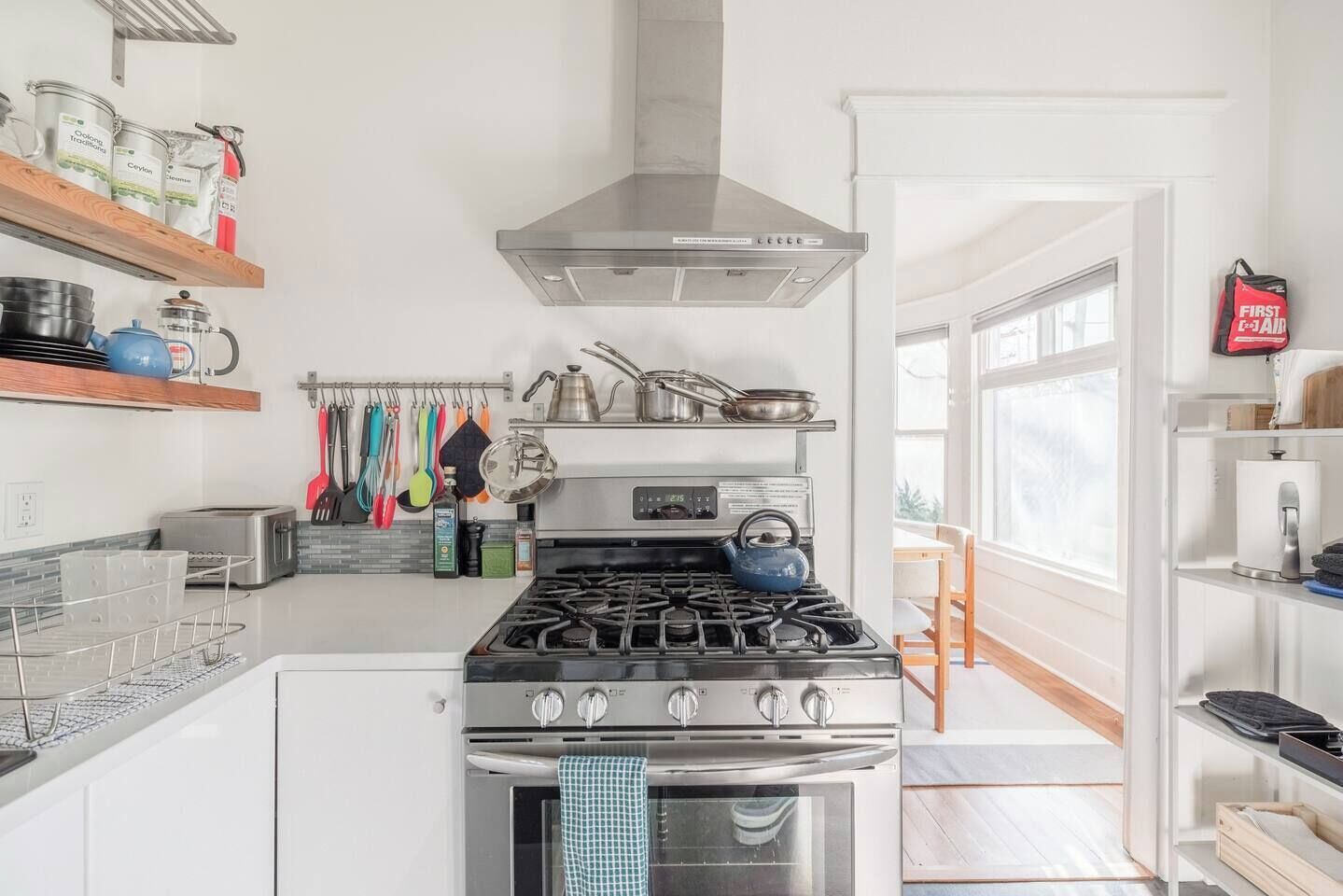 A modern kitchen with a gas stove, oven, and stainless steel appliances. There are shelves with kitchen utensils and a window providing natural light.