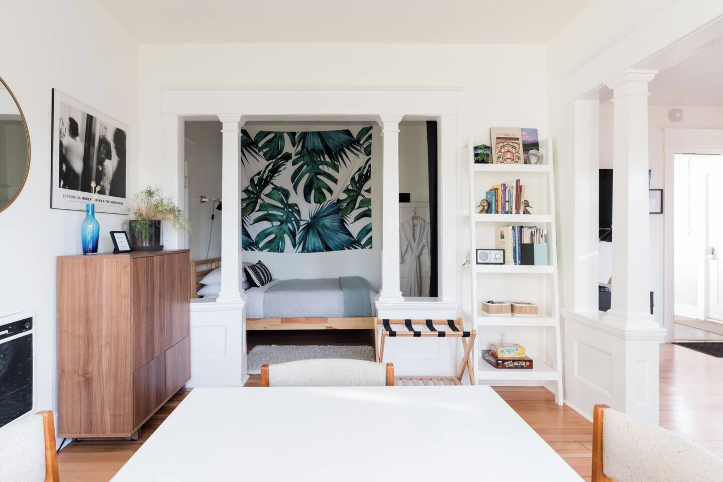 A modern bedroom with a bed, a side table, a bookshelf, and a large tropical leaf wall mural.