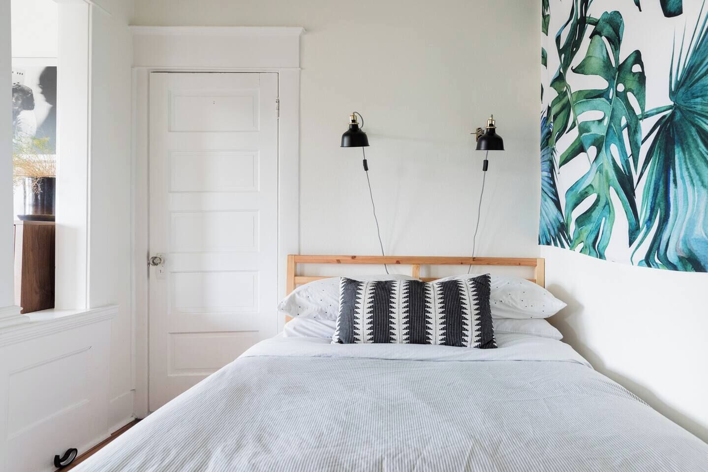 A bedroom with a bed, two wall-mounted lamps, a door, and a large tropical leaf painting.