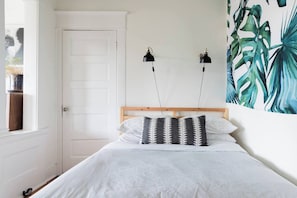 A bedroom with a bed, two wall-mounted lamps, a door, and a large tropical leaf painting. - Radiant, Low-Key Apartment (Seattle)