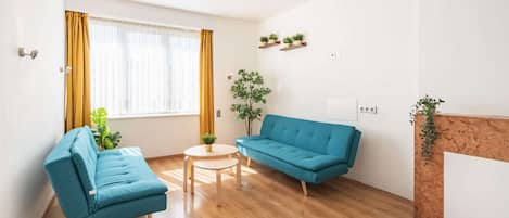 Ein modernes Wohnzimmer mit einem blauen Sofa, einem kleineren blauen Sessel, einem runden Couchtisch und einer Topfpflanze.