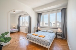 Un dormitorio con dos camas, ventanas amplias, un radiador y una planta en maceta.