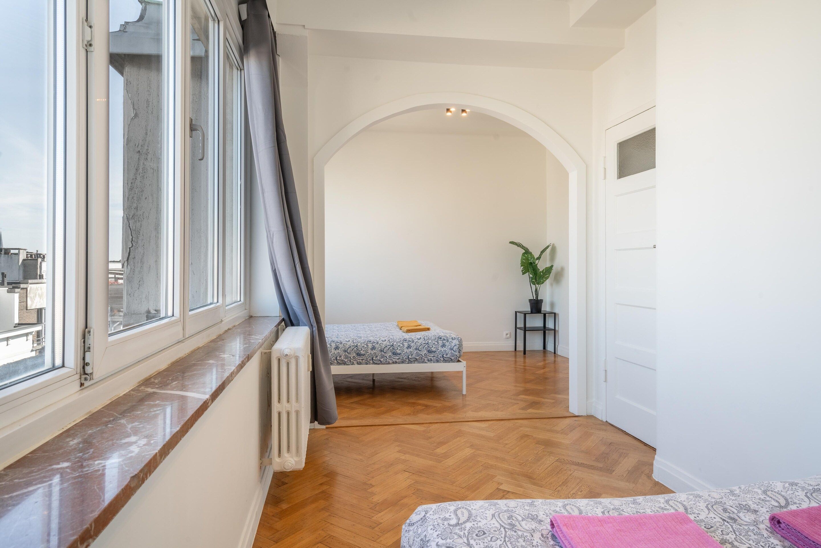 Une chambre à coucher avec un lit, un radiateur, une plante en pot et une vue sur la ville par la fenêtre.