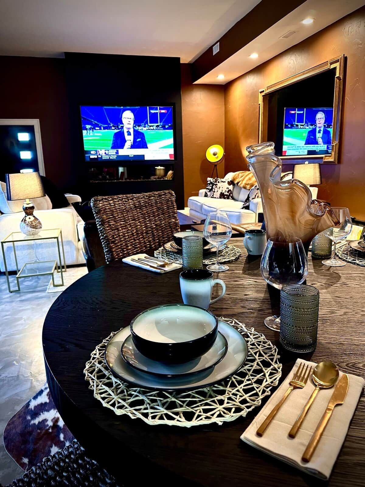 A dining area with a round table set for two, a pitcher, wine glasses, and a television displaying a sports event.