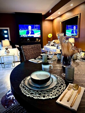A dining area with a round table set for two, a pitcher, wine glasses, and a television displaying a sports event. - Luxury & Wellness (Dayton)