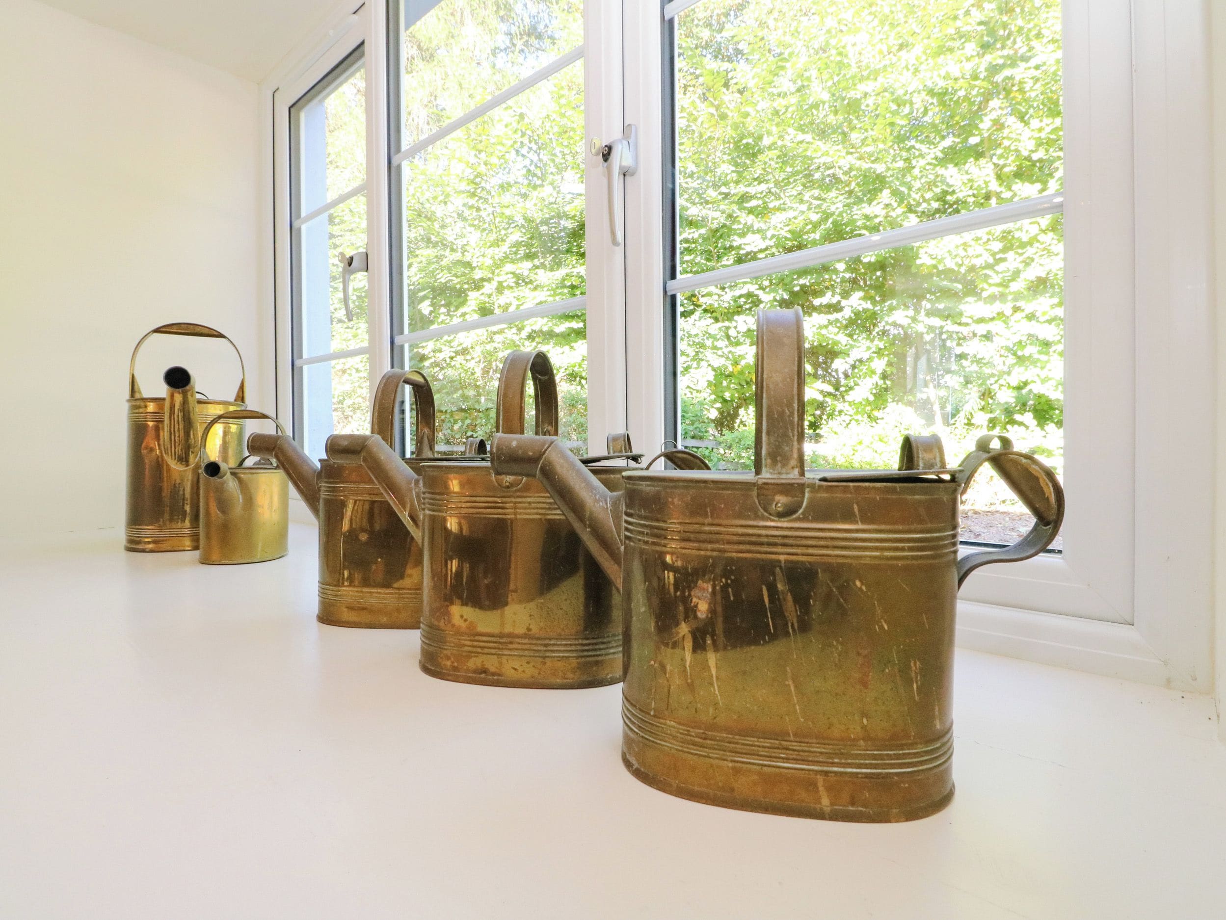 Four brass watering cans on a windowsill with a view of trees outside.
