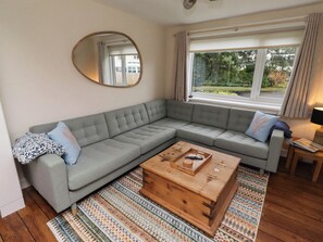 Ein Wohnzimmer mit grauem Ecksofa, einem Holztisch, einem Teppich mit Muster und einem großen Fenster mit Vorhängen.