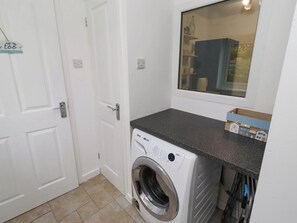 A laundry room with a front-loading washing machine, a countertop, and a window. - Bari House (Seahouses)