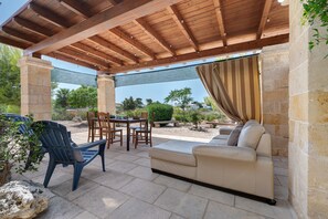 Patio esterno con pergola in legno, pareti in pietra, un tavolo con sedie e un divano color beige.