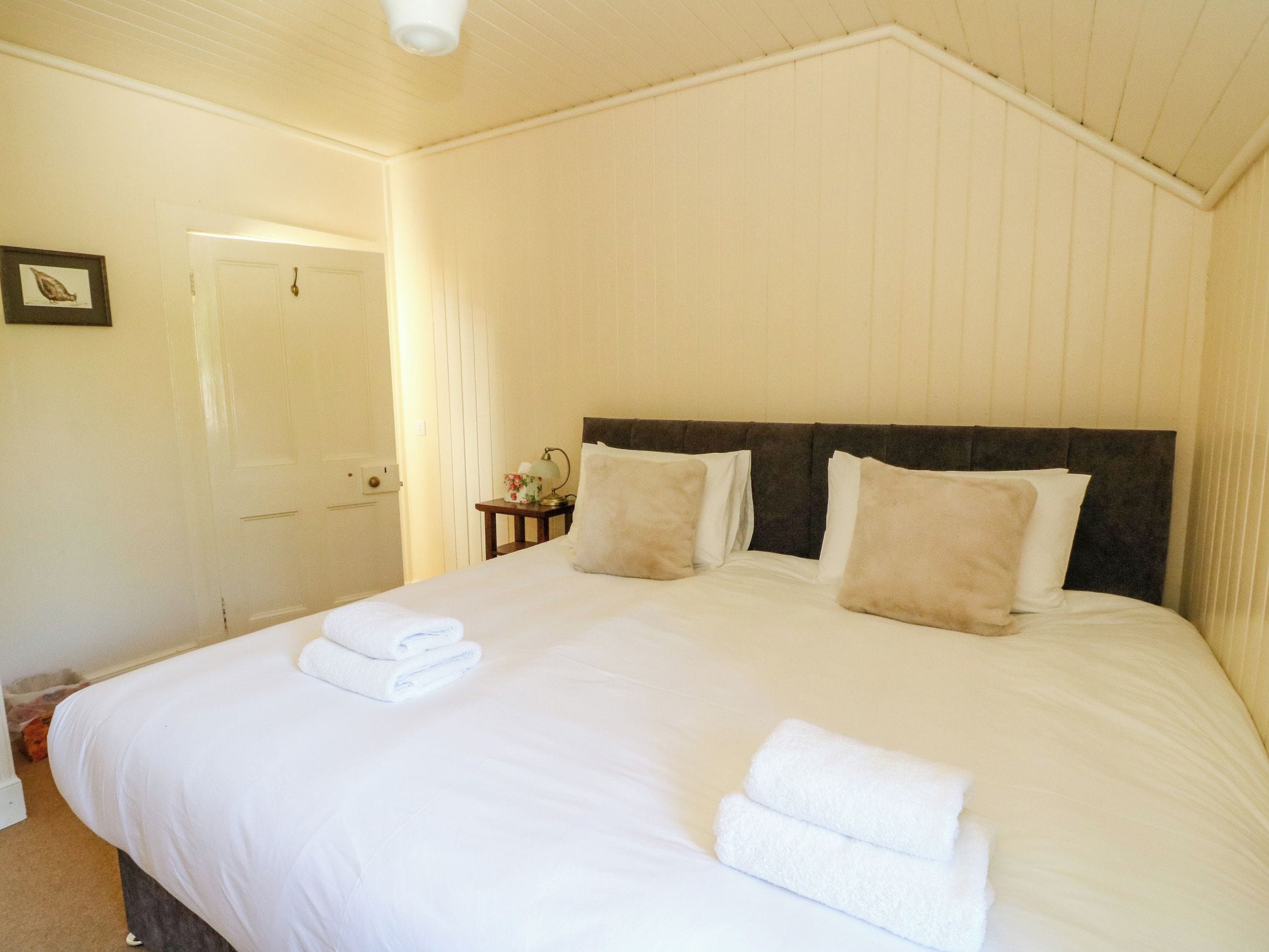 A neatly made bed with white linens and two pillows, a bedside table with a lamp, and a door in the background.
