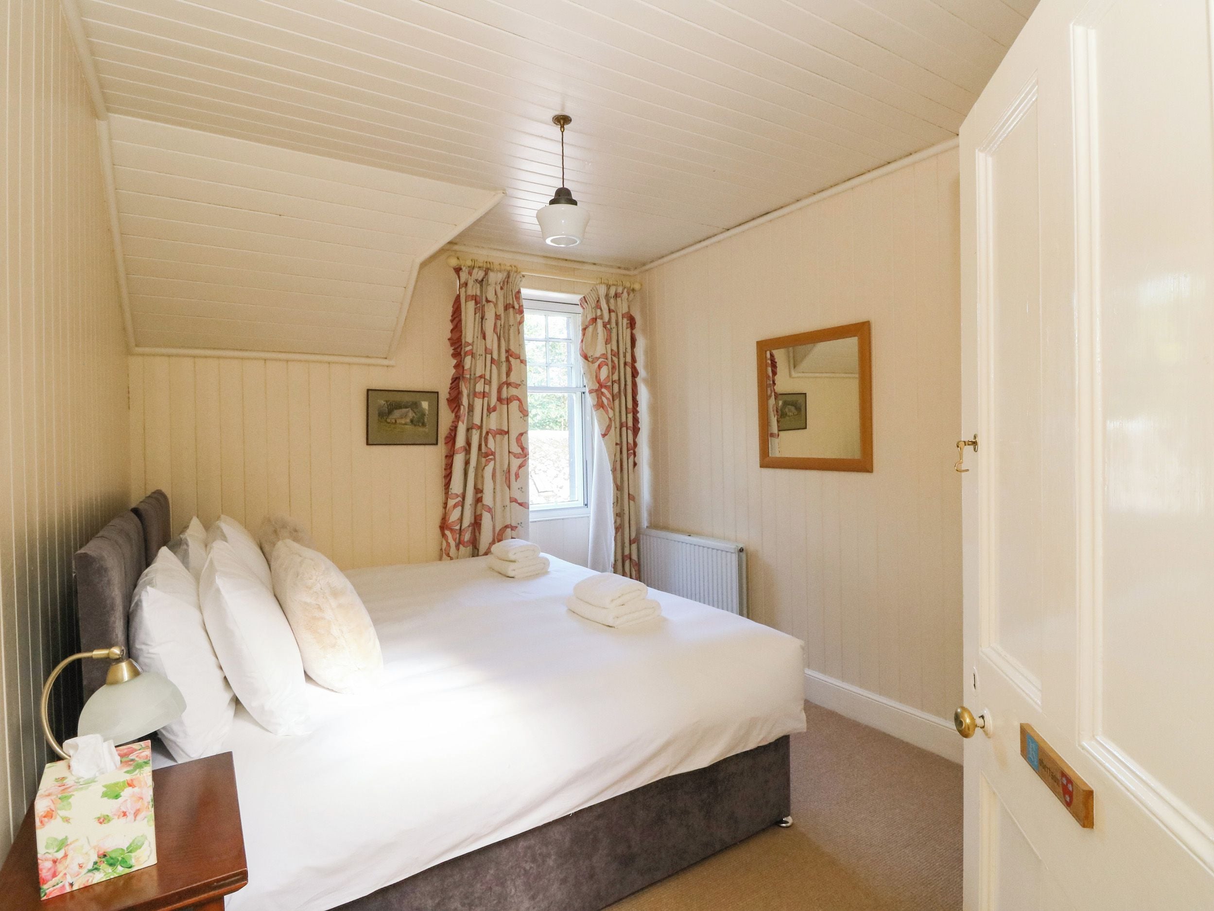 A bedroom with a bed, a bedside lamp, a radiator, a window with curtains, a mirror, and a door with a nameplate.