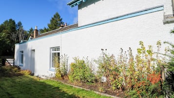 Un edificio blanco con detalles azules, una ventana y una puerta. Se observa un jardín con diversas plantas y una valla de madera al fondo.