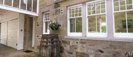 A stone building with a garage door, a wooden barrel, and a window with a hanging flower basket.