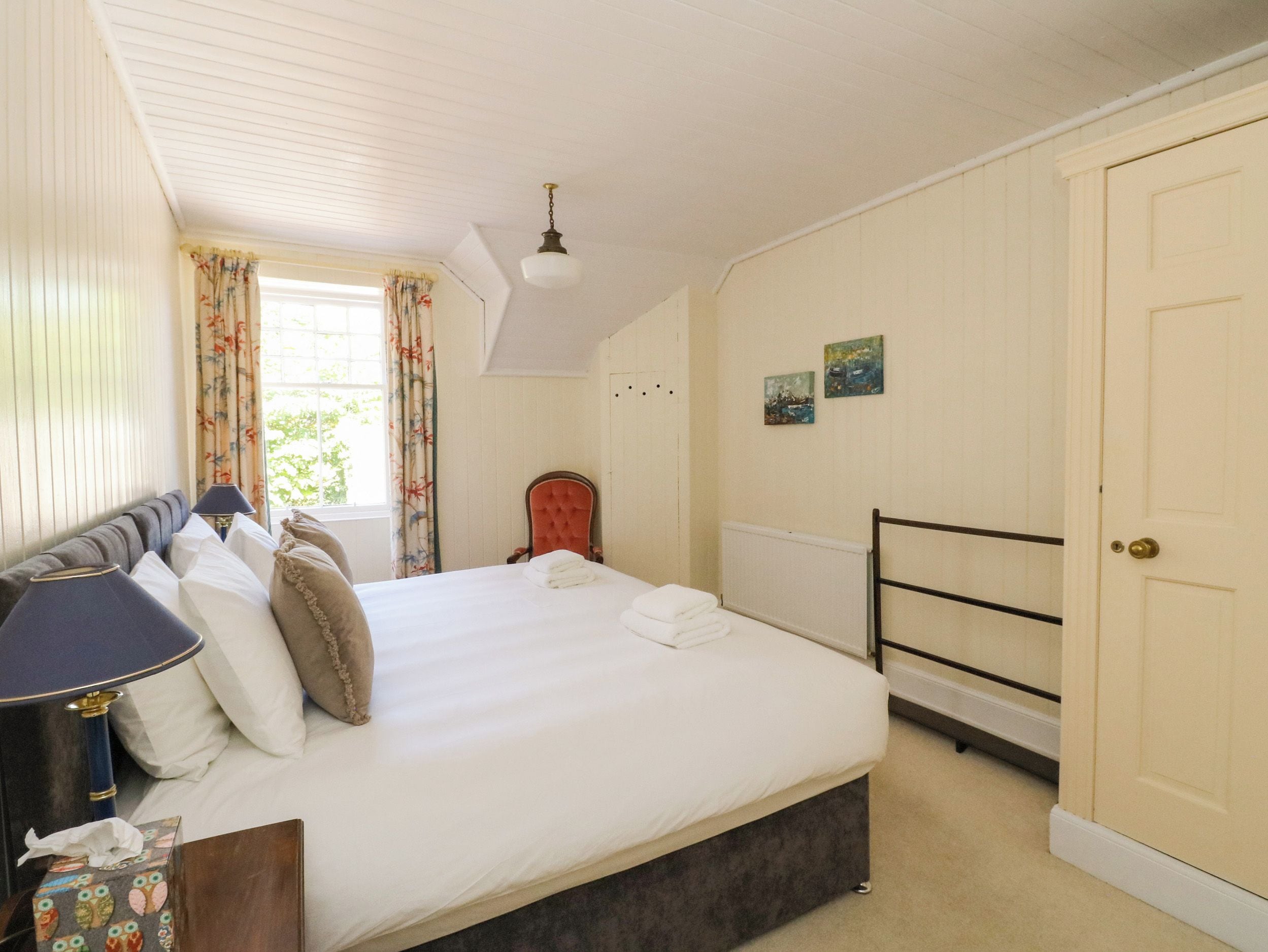 A bedroom with a bed, a chair, a bedside table, a lamp, a window with curtains, and a door.