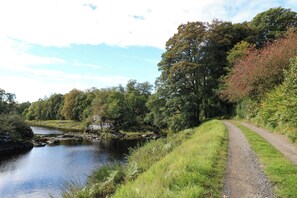Un sentier de gravier longe une rivière bordée d’arbres et surmontée d’une structure en pierre.