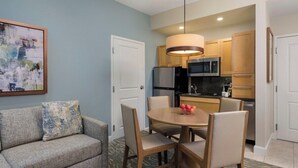 Un espacio habitable compacto con kitchenette, un comedor con mesa y sillas redondas, un sofá con un cojín estampado y un cuadro en la pared.