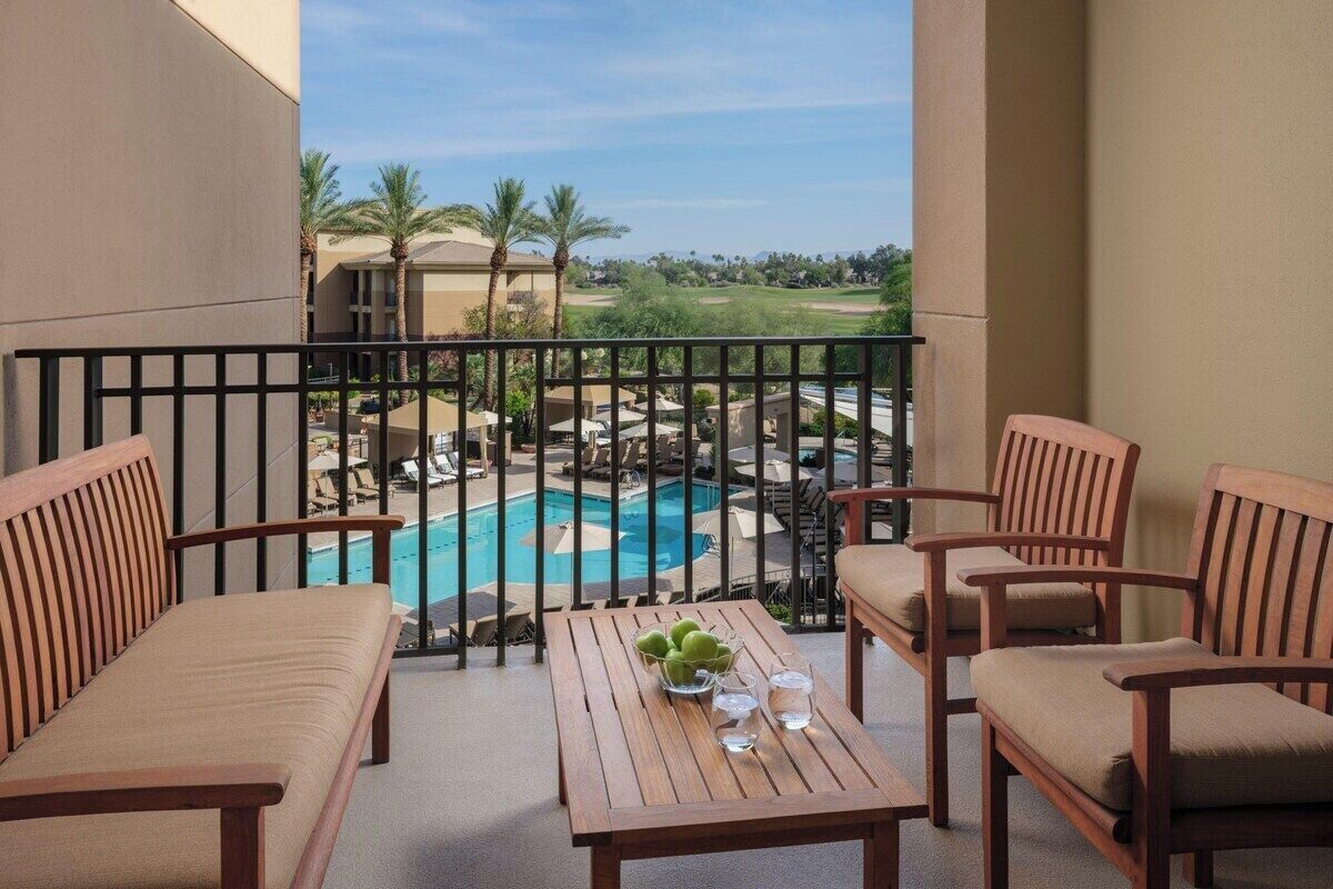Un balcón con vista a la piscina, muebles de madera y una mesa con frutas y vasos.