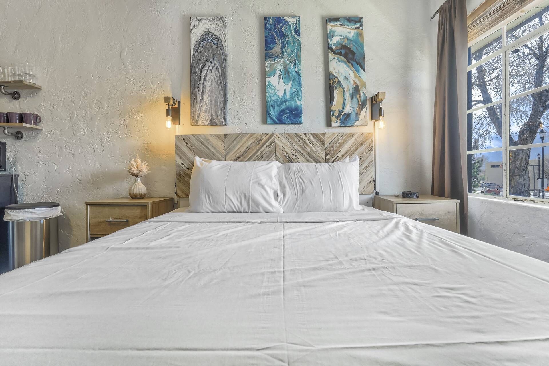Un dormitorio con una cama, dos mesitas de noche, una lámpara de noche, un ventanal con vistas y tres cuadros abstractos en la pared.