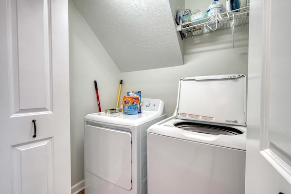 A laundry room with a washing machine and dryer, a shelf with cleaning supplies, and a door with a handle. - Cozy 3-bedroom entire home near wineries & waterfalls in Dahlonega, GA (Dahlonega)