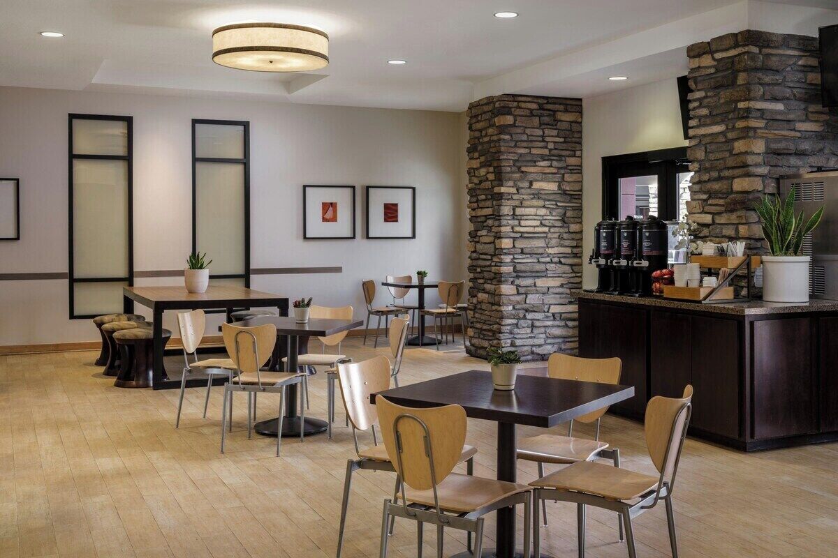 Uma área de refeições de hotel com várias mesas e cadeiras, uma parede de destaque em pedra e um bar com eletrodomésticos de cozinha.