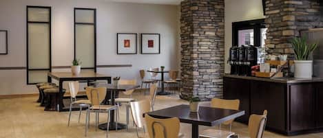 A hotel dining area with multiple tables and chairs, a stone accent wall, and a bar area with kitchen appliances.