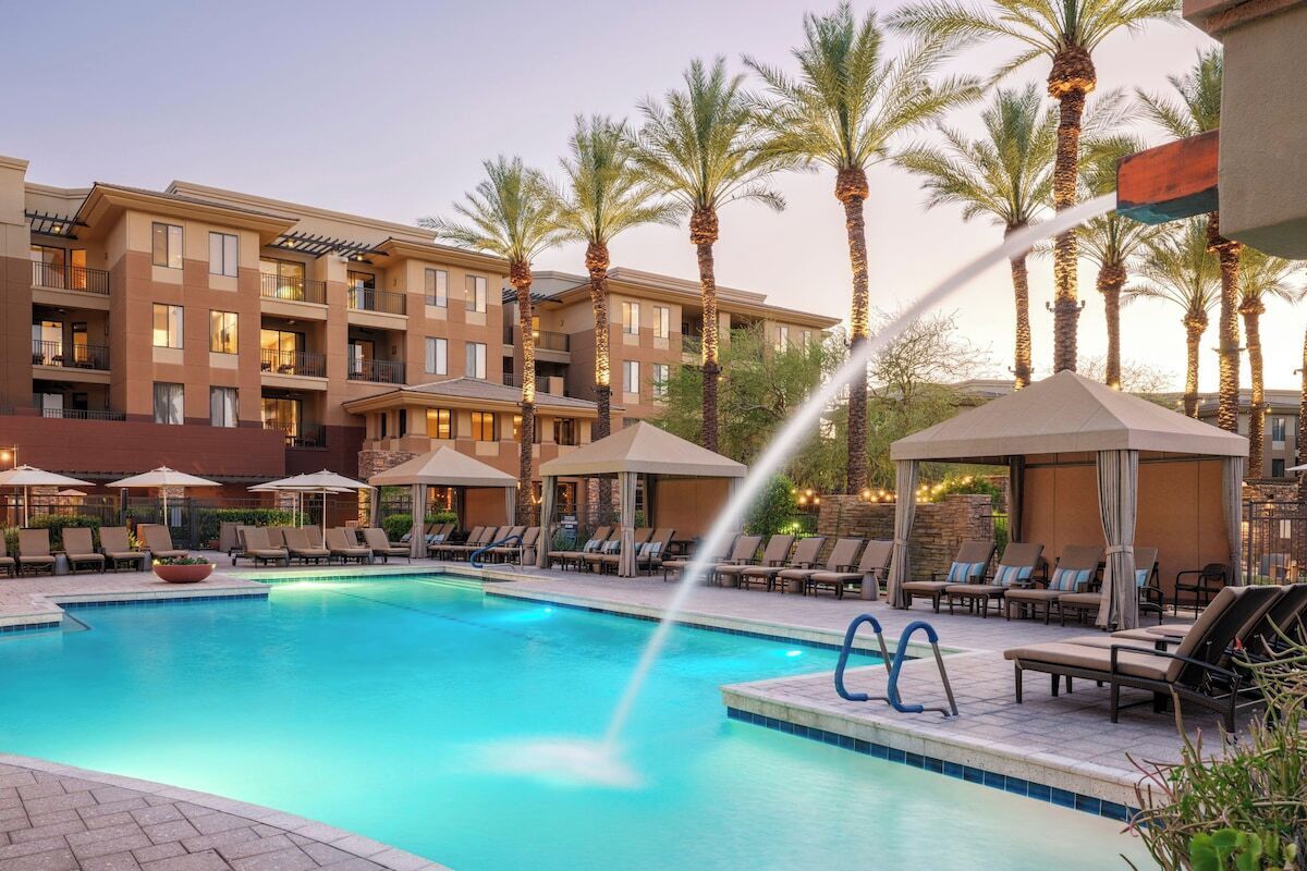 A hotel pool area with lounge chairs and umbrellas, surrounded by palm trees.