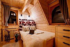 A cozy wooden cabin bedroom with a sloped ceiling, a bed with a laptop, a window with curtains, and a small table with a vase of flowers. - Comfortable Mountain-style House With a SPA Area (Poronin)