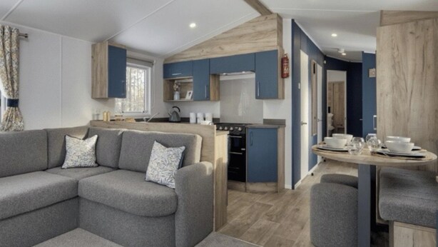 A modern kitchen with blue cabinets, a dining table with chairs, and a grey sofa with patterned cushions. - Thurston Manor, 3 Bedroom Caravan (Dunbar)