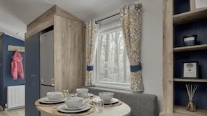A dining area with a round table set for four, a window with patterned curtains, and a bookshelf with a framed picture. - Thurston Manor, 3 Bedroom Caravan (Dunbar)