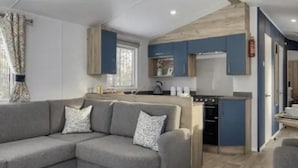 A modern living room with a grey sectional sofa and a kitchen area with blue cabinets and a wooden ceiling. - Thurston Manor, 3 Bedroom Caravan (Dunbar)
