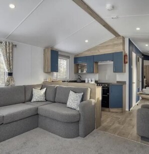 A modern living room with a grey sectional sofa and a kitchen area with blue cabinets and a wooden ceiling. - Thurston Manor, 3 Bedroom Caravan (Dunbar)
