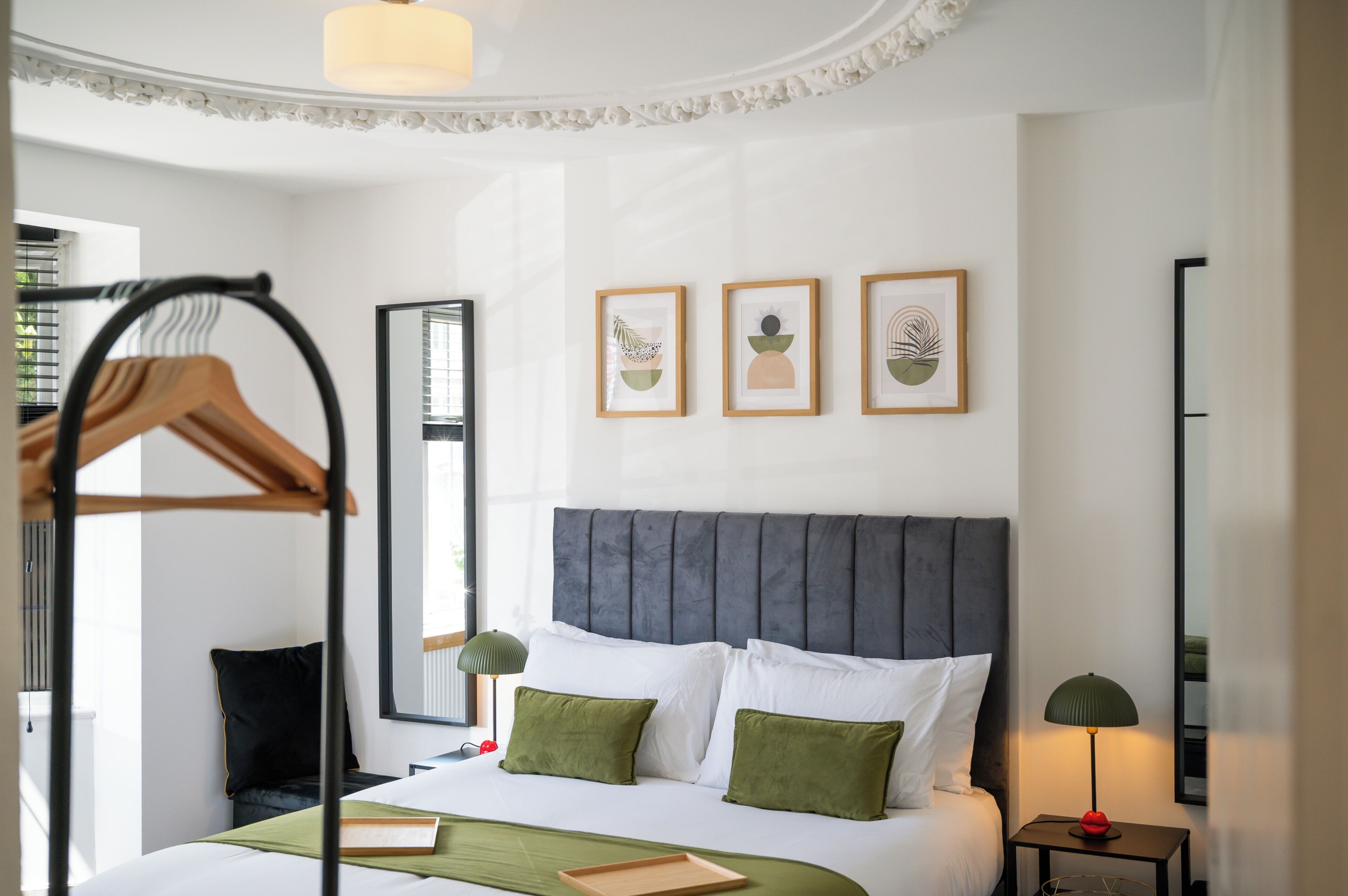 Une chambre d’hôtel avec un lit, une table de chevet, une lampe, un miroir et trois œuvres d’art encadrées accrochées au mur.
