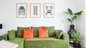 Un canapé vert agrémenté de coussins orange, un vase noir contenant une plante et trois œuvres d’art abstraites encadrées accrochées au mur.