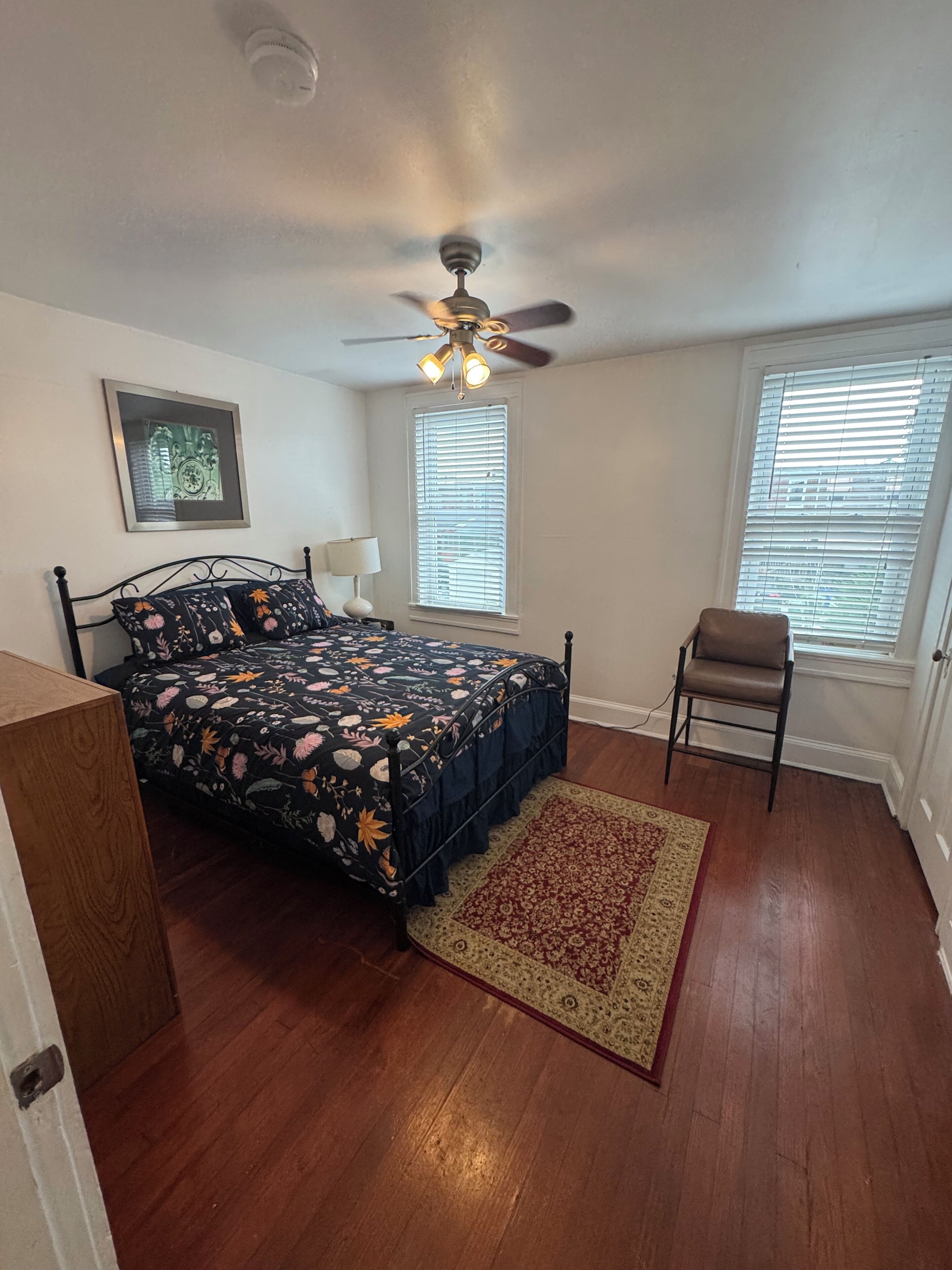 A bedroom with a bed, a chair, a ceiling fan, a window with blinds, and a framed picture on the wall.