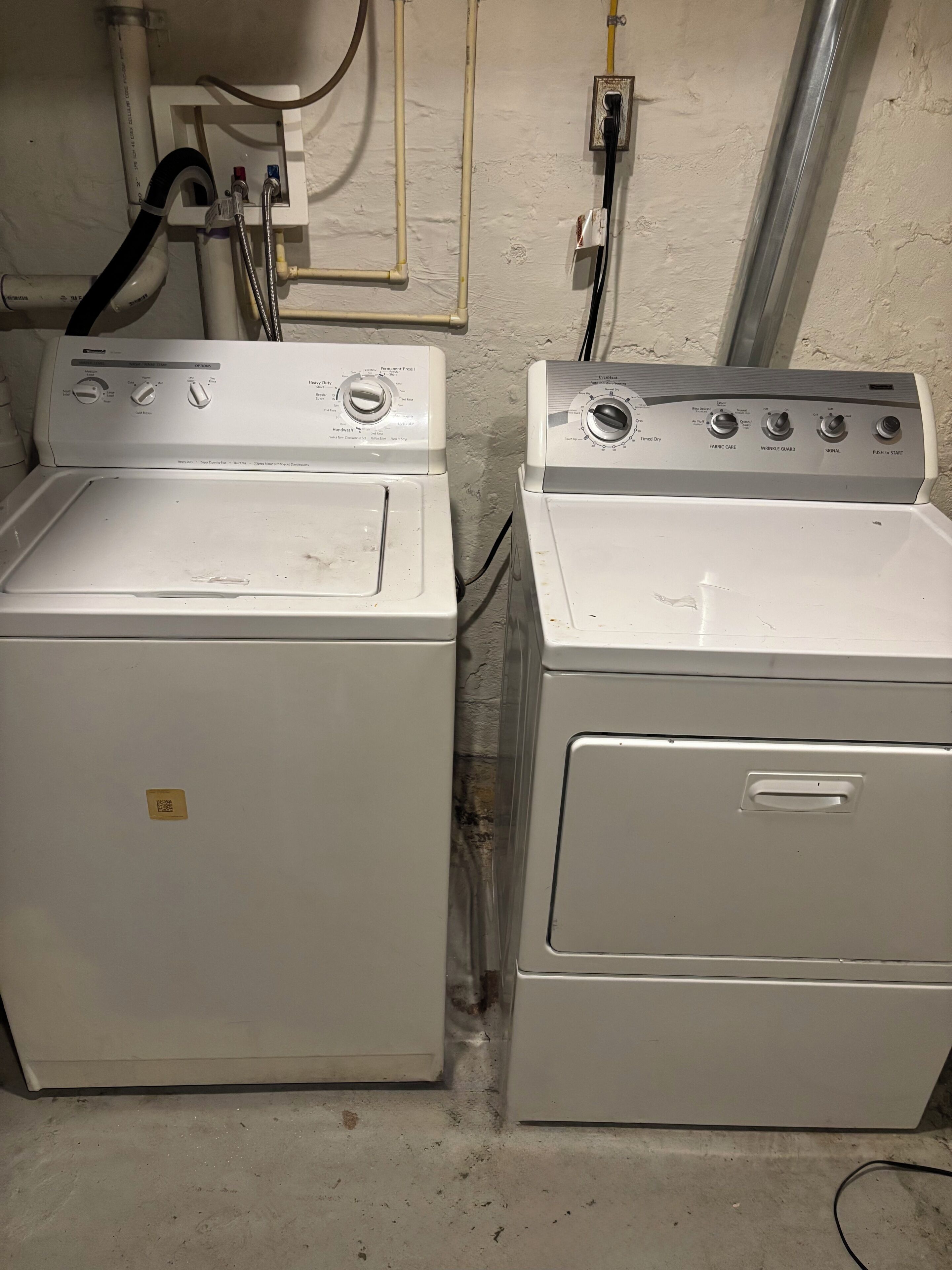 Deux machines à laver à chargement frontal, placées côte à côte, avec des panneaux de commande et des tuyaux visibles.