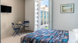 Un dormitori amb un llit, una televisió penjada a la paret, un balcó amb vista a edificis i una taula petita amb cadires.